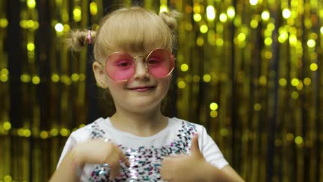 Child-show-thumbs-up.-Smiling,-looking-at-camera.-Girl-posing-on-background-with-foil-golden-curtain