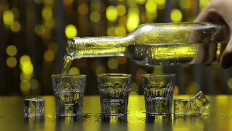 Barmann-Gießt-Gefrorenen-Wodka-Aus-Der-Flasche-In-Ein-Schnapsglas.-Eiswürfel-Vor-Goldglänzendem-Partyhintergrund