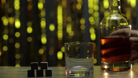Gießen-Von-Goldenem-Whiskey,-Cognac-Oder-Brandy-Aus-Der-Flasche-In-Ein-Glas-Mit-Steinen,-Eissteinen-Auf-Dem-Tisch