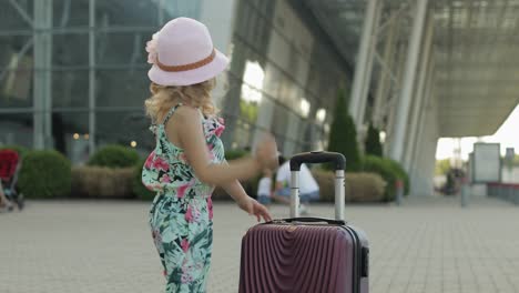 Child-girl-tourist-with-suitcase-bag-near-airport.-Kid-with-luggage-dances,-rejoices,-show-thumbs-up