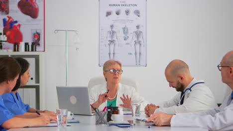 Elderly-specialist-woman-doctor-sitting-on-conference-desk-talking-with-team