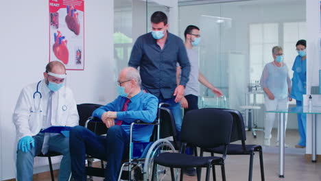 Anciano-Inválido-Con-Mascarilla-Discutiendo-Con-El-Médico-En-La-Sala-De-Espera-Del-Hospital