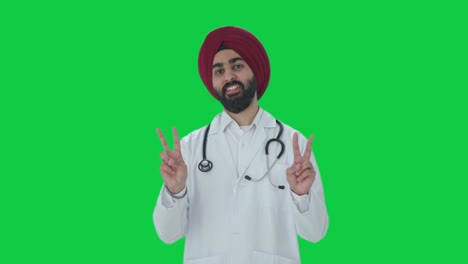 Fröhlicher-Indischer-Sikh-Arzt-Zeigt-Siegeszeichen-Auf-Grünem-Bildschirm