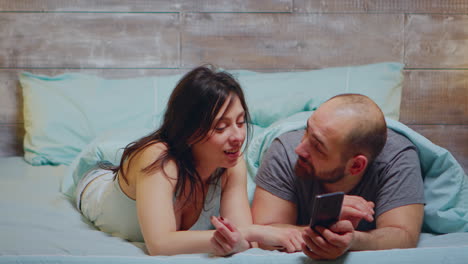 Wife-in-pajamas-laughing-while-her-husband-is-showing-something-on-phone