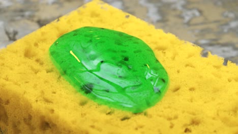 Close-up-shot-of-green-dish-soap-being-squeezed-onto-a-yellow-sponge-placed-on-a-rusted-metal-kitchen-surface.-Dishwashing-liquid-detergent-is-poured-on-a-dish-scrub-in-clean-macro-shot.