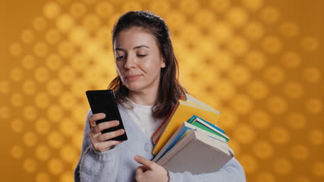 Smiling-woman-reading-messages-on-smartphone-while-holding-stack-of-books