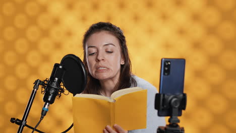 Influencer-filming-reading-book-with-smartphone-on-tripod,-studio-background
