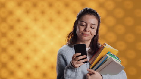 Smiling-woman-reading-messages-on-smartphone-while-holding-stack-of-books