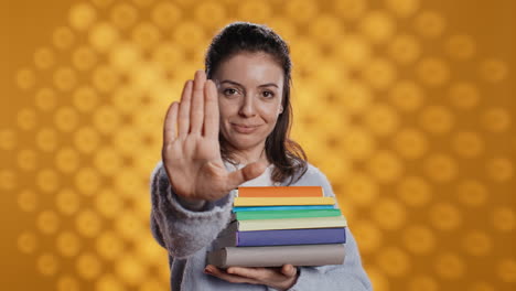 Portrait-of-stern-woman-holding-stack-of-books-doing-stop-sign-gesturing