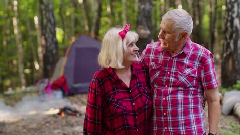 Senior-elderly-grandmother-grandfather-tourists-resting-at-camping-in-wood-over-bonfire-at-camping