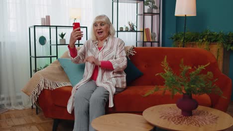 Glückliche-ältere-Großmutter-Hält-Smartphone-Und-Schaut-Sich-Mobile-Videoanrufe-Online-An-Und-Macht-Selfie-Zu-Hause