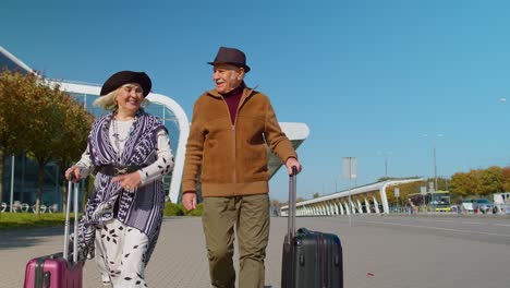 Elderly-husband-wife-retirees-tourists-go-to-airport-terminal-for-boarding-with-luggage-on-wheels