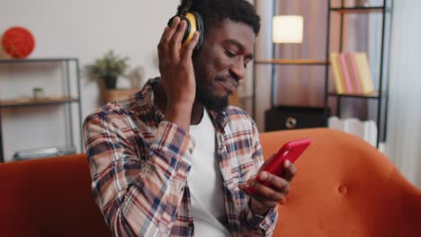 African-American-young-man-in-headphones-listening-music-dancing,-singing-in-living-room-at-home