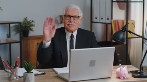 Senior-business-man-waves-hand-palm-in-hi-gesture-greeting-welcomes-someone-webinar-at-home-office