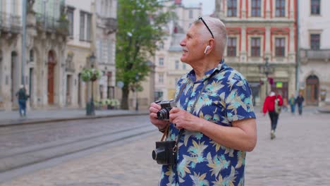 Senior-tourist-with-retro-photo-camera-listening-to-music-on-earphones-dancing-outdoors-having-fun