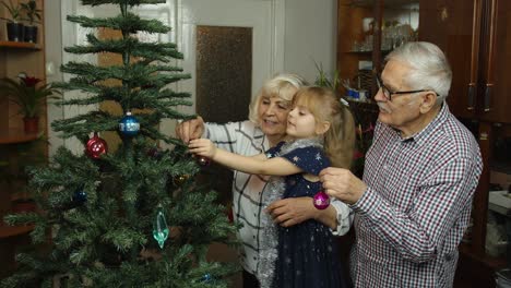 Child-girl-with-senior-grandparents-family-decorating-artificial-Christmas-tree,-New-Year-holidays