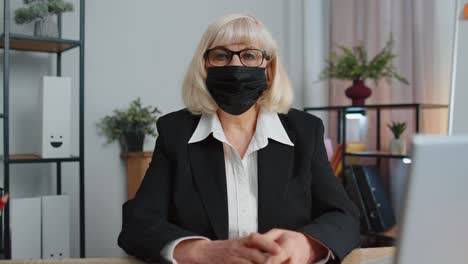 Senior-employee-wears-face-mask-works-alone-from-home-office-sits-at-desk-looking-at-camera