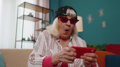 Worried-funny-grandmother-woman-enthusiastically-playing-racing-video-games-on-mobile-phone-at-home