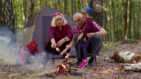 Happy-senior-man-woman-with-grandchild-talking-cooking-frying-sausages-over-campfire-in-evening-wood