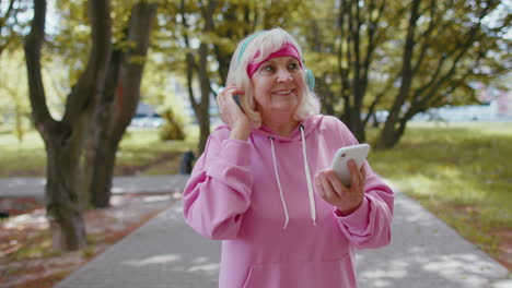 Athletic-senior-sport-runner-woman-wearing-earphones-listening-music-from-smartphone-in-morning-park
