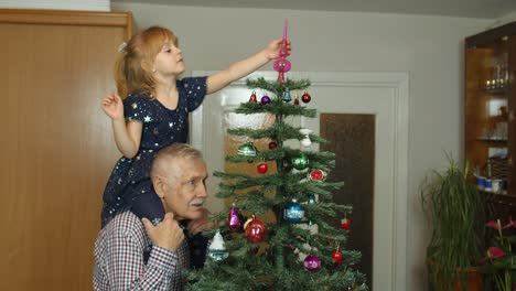Kid-girl-with-senior-grandfather-decorating-artificial-Christmas-tree-with-ornaments-toys-at-home