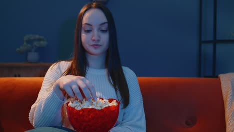 Woman-sitting-on-sofa-alone,-eating-popcorn-and-watching-interesting-tv-serial,-horror-film-at-home
