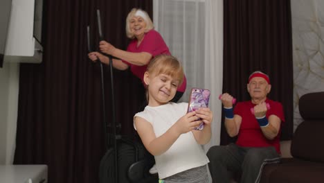 Senior-couple-using-orbitrek-doing-dumbbells-exercises,-child-girl-making-selfie-on-mobile-phone