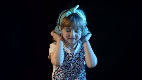 Stylish-teenager-kid-girl-standing-among-neon-lights-listening-to-music-in-headphones,-dancing