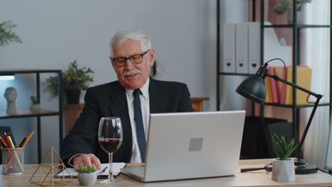 Senior-man-freelancer-drinking-wine-after-working-on-laptop-computer-pc-sitting-at-desk-at-office