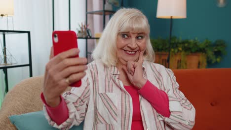 Glückliche-ältere-Großmutter-Hält-Smartphone-Und-Schaut-Sich-Mobile-Videoanrufe-Online-An-Und-Macht-Selfie-Zu-Hause