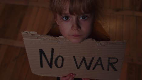 Afraid-homeless-toddler-girl-sitting-holding-inscription-No-War,-hiding-from-bombing-attack-at-home