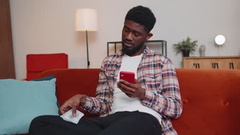 Sad-poor-african-american-man-holding-cash-money-and-mobile-phone-calculate-domestic-bills-at-home