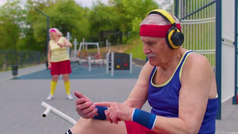 Senior-man-after-sport-training-listening-music-from-mobile-phone-wearing-headphones-on-playground