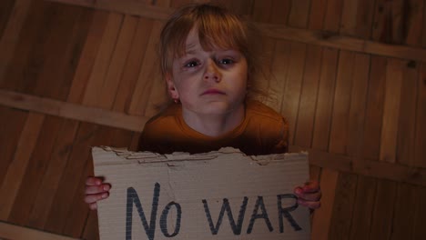 Scared-homeless-toddler-girl-sitting-holding-inscription-No-War,-hiding-from-bombing-attack-at-home