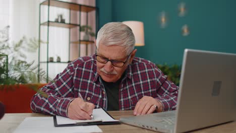 Älterer-Mann,-Der-Am-Laptop-Arbeitet-Und-Sich-Notizen-Auf-Einem-Blatt-Papier-Macht,-Verärgert-über-Schlechte-Ergebnisse