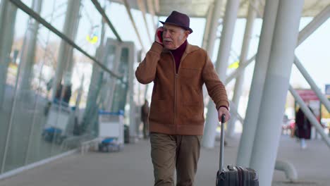 Senior-tourist-grandfather-man-walking-on-international-airport-hall,-using-mobile-phone,-talking