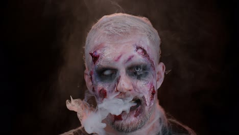 Sinister-man-horrible-scary-Halloween-zombie-looking-ominous-at-camera-blows-smoke-from-nose,-mouth