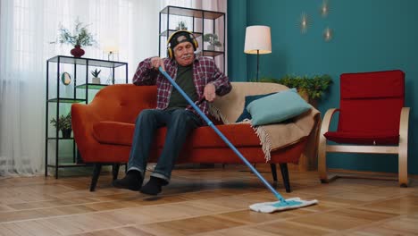 Happy-senior-grandfather-housekeeper-is-mopping-floor-in-living-home-room,-listening-music,-dancing