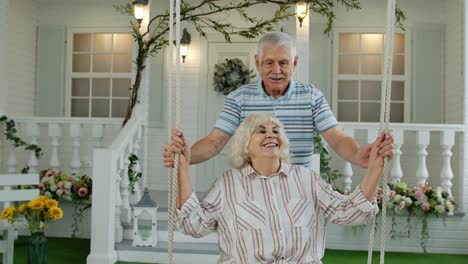 Senior-couple-together-in-front-yard-at-home.-Man-swinging-woman.-Happy-mature-retired-family