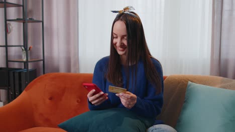 Woman-sitting-at-home-using-credit-bank-card-and-smartphone-while-transferring-money-online-shopping
