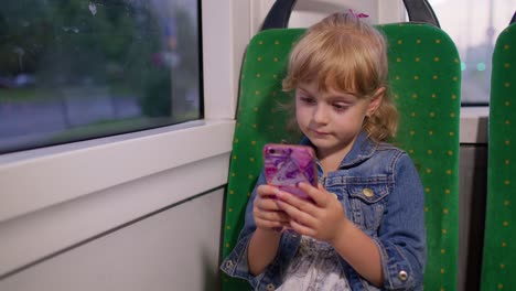 Smiling-attractive-child-girl-kid-in-city-train,-tram-or-bus-using-smartphone-chatting-with-friends