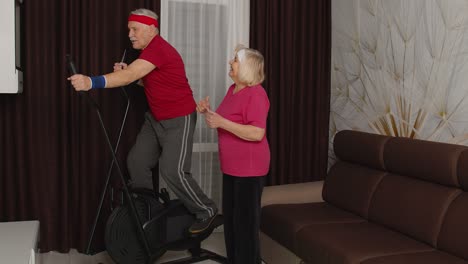 Active-senior-mature-grandfather-man-in-sportswear-using-orbitrek,-grandma-encourages-him-at-home