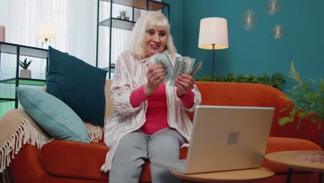 Satisfied-cheerful-senior-grandmother-freelancer-woman-pressing-button-on-laptop-and-counting-money