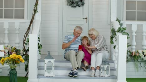 Älteres-Paar-Sitzt-Mit-Enkelin-Auf-Der-Veranda-Zu-Hause.-Nutzung-Des-Mobiltelefons-Für-Online-Videoanrufe