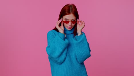 Sincere-cool-cheerful-redhead-girl-in-blue-sweater-wearing-sunglasses,-charming-smile-on-pink-wall