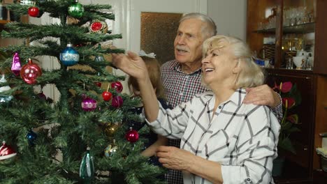 Child-girl-with-senior-grandparents-family-decorating-artificial-Christmas-tree,-New-Year-holidays