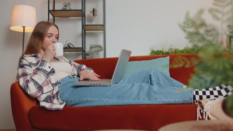 Young-woman-using-laptop-computer-lying-on-sofa-at-home-working,-online-shopping-from-home-office