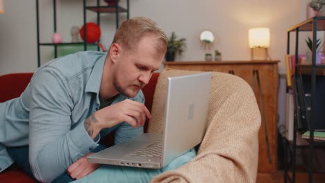 Hombre-Adulto-Usando-Una-Computadora-Portátil-Tumbado-En-Un-Sofá-En-Casa-Trabajando,-Comprando-En-Línea,-Viendo-Películas