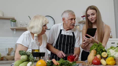 Mature-grandparents-couple-cutting-vegetables-for-salad,-listening-recipe-from-girl-with-tablet