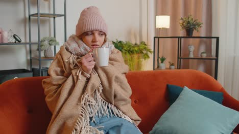 Young-sick-woman-wear-hat-wrapped-in-plaid-sit-alone-shivering-from-cold-on-sofa-drinking-hot-tea